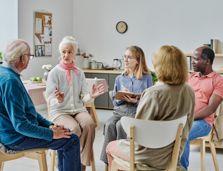 Un grupo de personas en círculo escucha a una persona mayor hablando mientras otra persona anota datos.