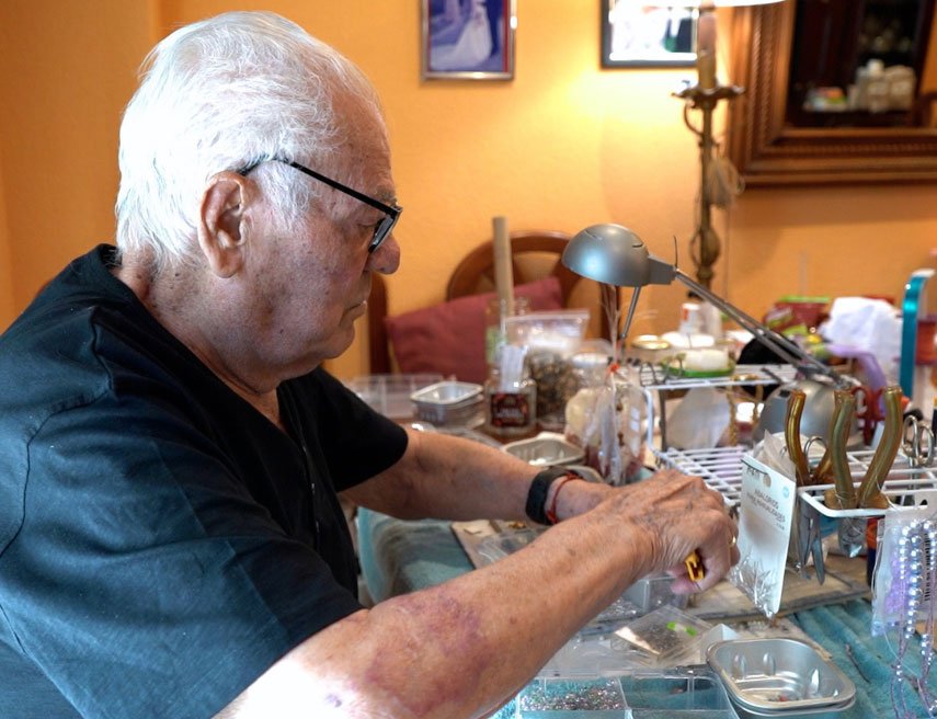 La imagen muestra a un hombre mayor sentado en una mesa realizando una actividad artesanal, posiblemente relacionada con la creación de bisutería o manualidades, ya que se observan herramientas, bandejas con abalorios y materiales clasificados. Está concentrado, con la mirada dirigida hacia su trabajo, en un entorno cálido y hogareño, iluminado por una lámpara de escritorio. La escena transmite una sensación de tranquilidad y dedicación, y podría estar relacionada con un proyecto de envejecimiento activo, soledad no deseada o actividades ocupacionales en el hogar.