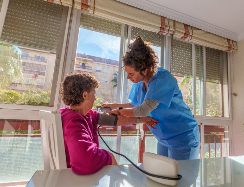 Una mujer es atendida por una persona cuidadora que le está tomando la tensión en su casa.