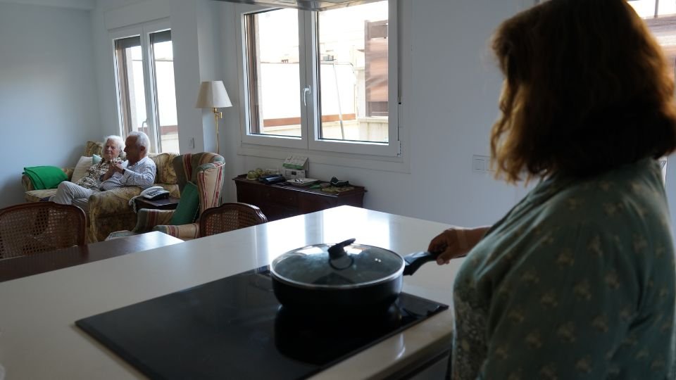 Teresa cocinando con Isidro y Araceli al fondo, viendo la televisión.