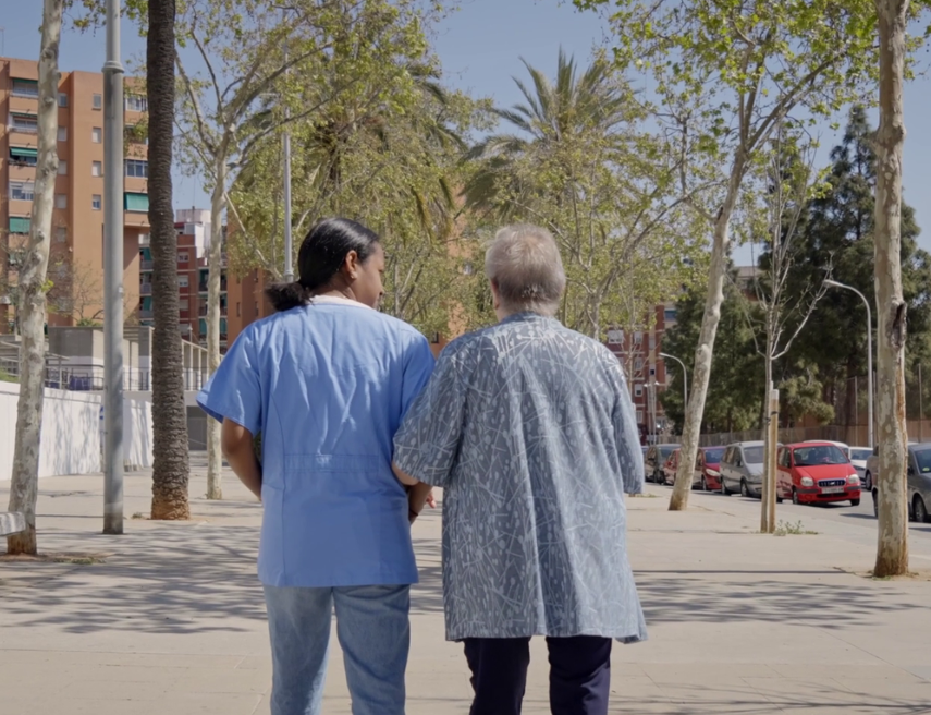 Dos mujeres, una mayor y otra cuidadora, caminan de espaldas por una calle cogidas de la mano.